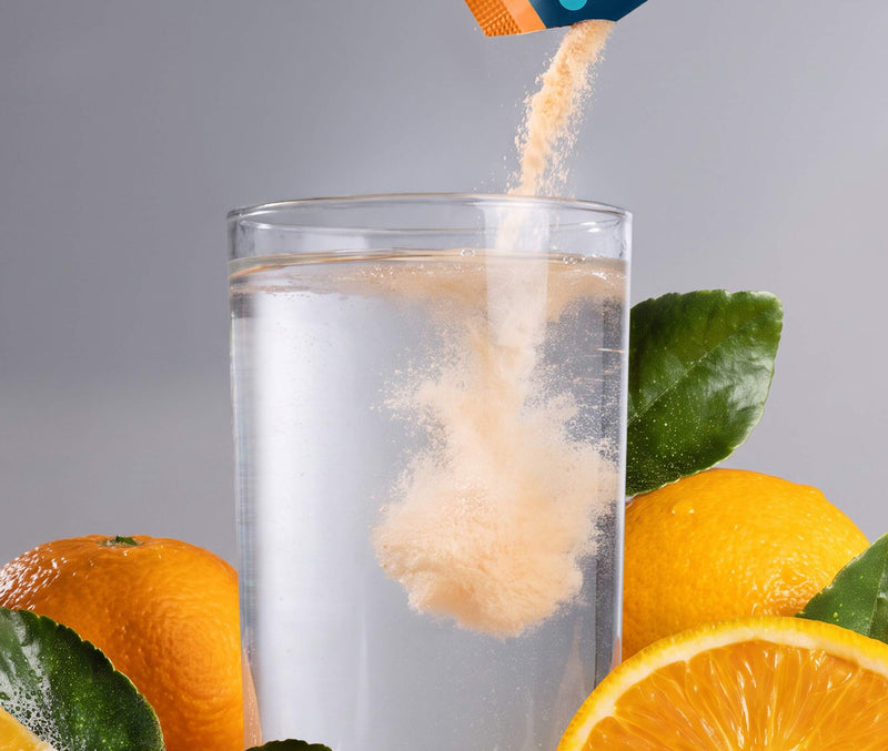 A close-up of a powder supplement being poured from a blue and orange sachet into a clear glass of water, surrounded by fresh oranges and green leaves on a gray background.
