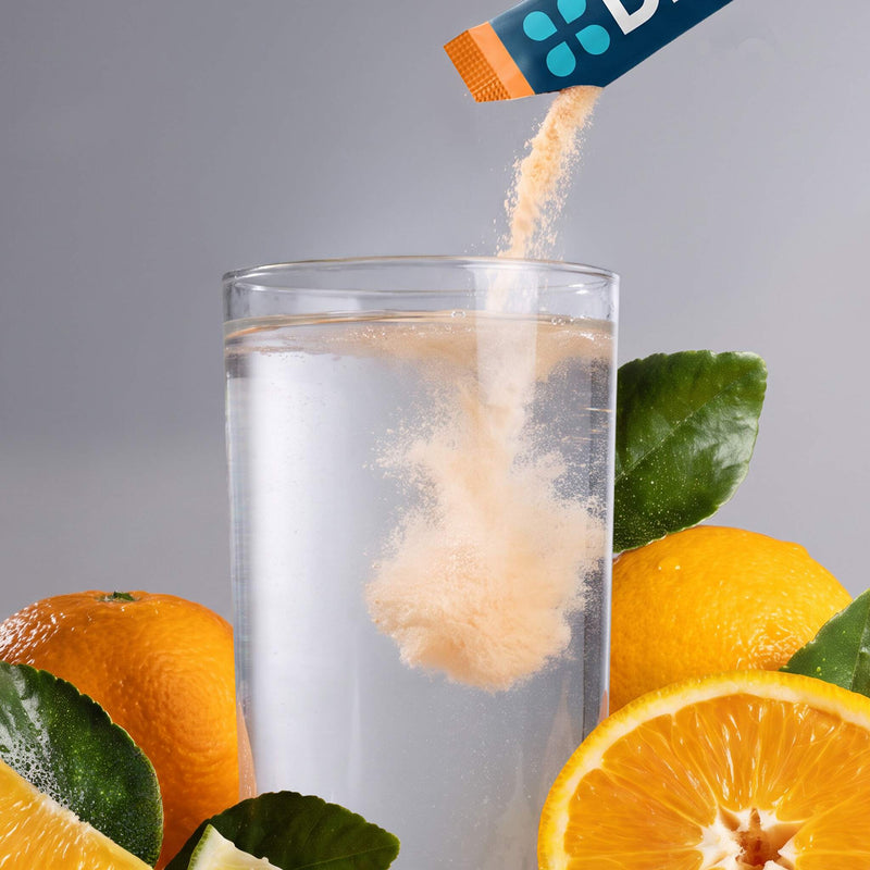 A close-up of a powder supplement being poured from a blue and orange sachet into a clear glass of water, surrounded by fresh oranges and green leaves on a gray background.