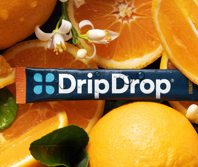 A DripDrop orange-flavored stick placed across a tightly arranged background of whole oranges, orange slices, and white citrus blossoms. 