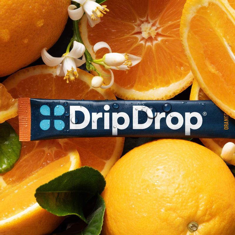 A DripDrop orange-flavored stick placed across a tightly arranged background of whole oranges, orange slices, and white citrus blossoms. 