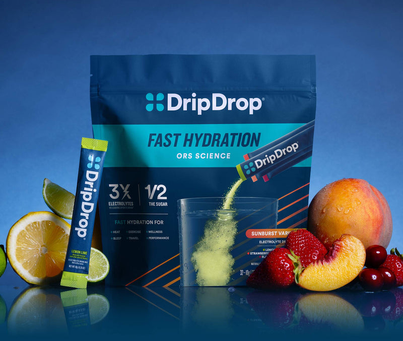 DripDrop Fast Hydration ORS Science powder pouch in blue packaging with a single stick pack beside it, shown with fruit and citrus slices on a blue studio background. The powder is being poured into a clear cup.