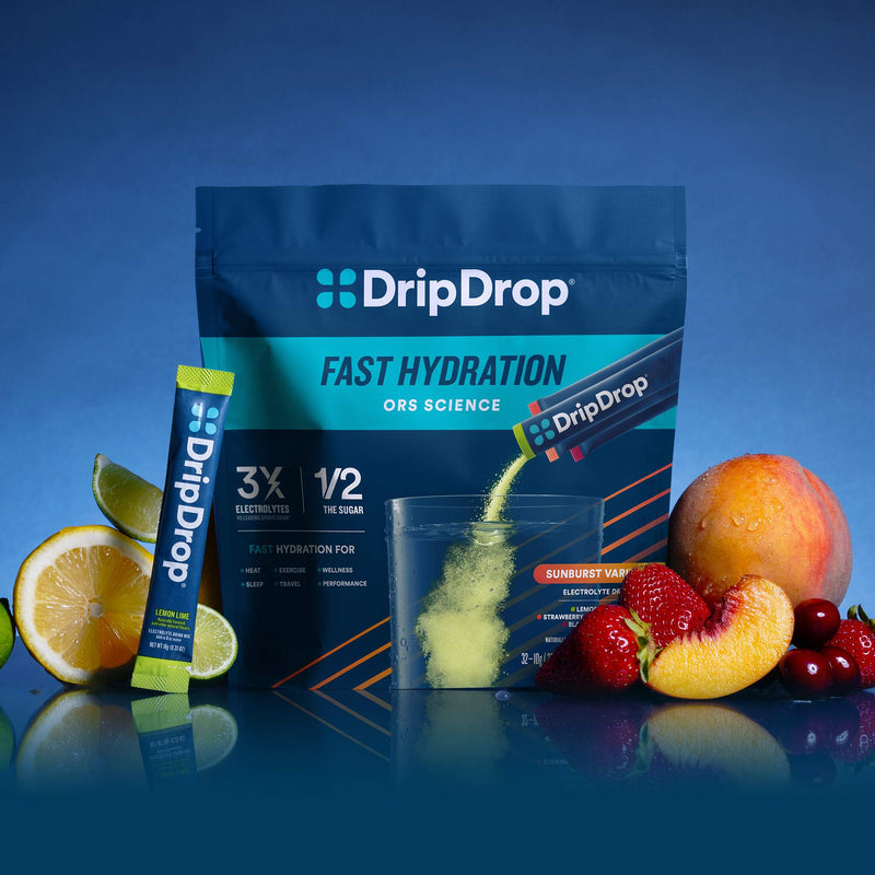 DripDrop Fast Hydration ORS Science powder pouch in blue packaging with a single stick pack beside it, shown with fruit and citrus slices on a blue studio background. The powder is being poured into a clear cup.
