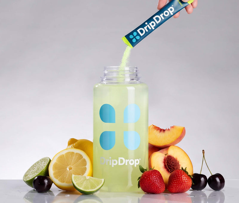 A hand pours DripDrop powder from a blue and lime sachet into a clear bottle labeled DripDrop, surrounded by fresh fruit slices including lemons, limes, strawberries, and peaches on a reflective white surface against a light gray background.