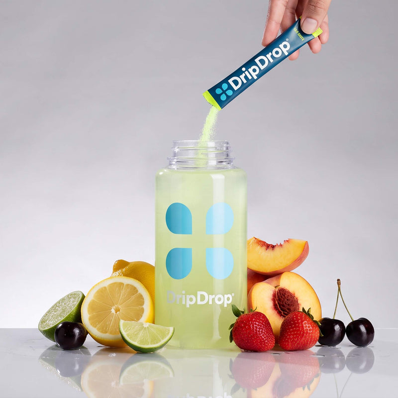 A hand pours DripDrop powder from a blue and lime sachet into a clear bottle labeled DripDrop, surrounded by fresh fruit slices including lemons, limes, strawberries, and peaches on a reflective white surface against a light gray background.