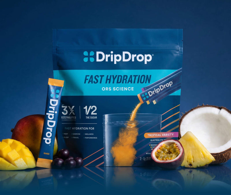 A stand-up pouch of DripDrop Fast Hydration ORS Science powder, shown with a single stick pack, a clear cup, and fruit slices and pieces including mango, coconut, pineapple, and passion fruit on a dark blue background.