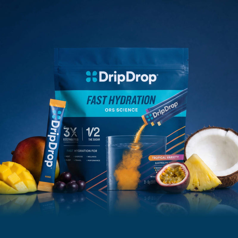 A stand-up pouch of DripDrop Fast Hydration ORS Science powder, shown with a single stick pack, a clear cup, and fruit slices and pieces including mango, coconut, pineapple, and passion fruit on a dark blue background.