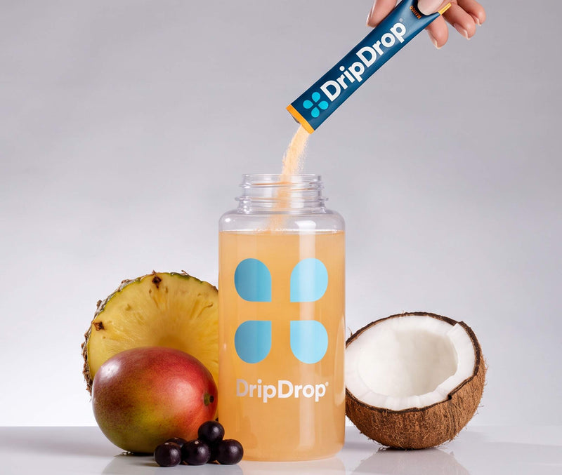 A hand pours powder from a DripDrop packet into a clear bottle filled with orange drink, styled with pineapple, mango, coconut, and berries on a light gray background.