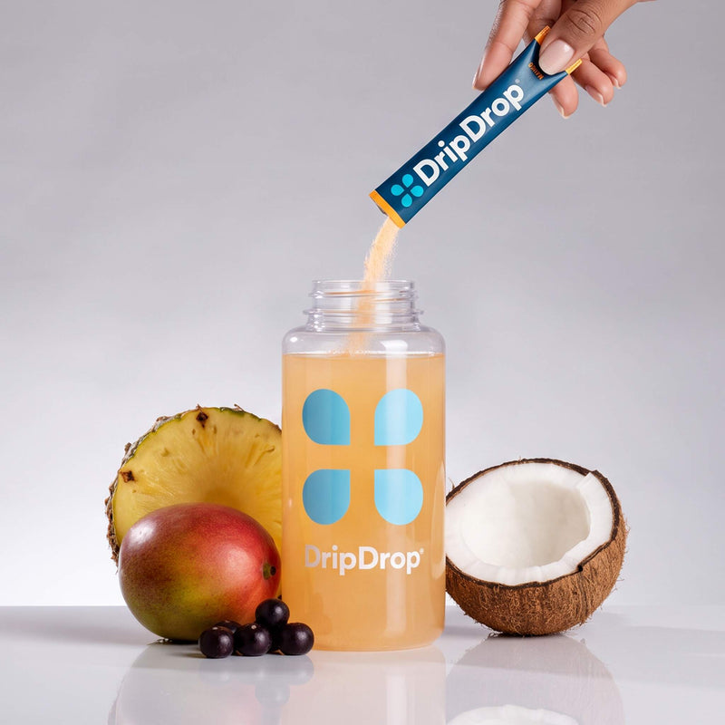 A hand pours powder from a DripDrop packet into a clear bottle filled with orange drink, styled with pineapple, mango, coconut, and berries on a light gray background.