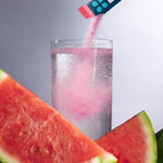 A clear glass filled with a clear liquid, with pink powder being poured into it from a small packet. Large watermelon slices frame the foreground against a soft gray studio background.