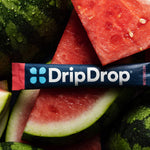 A DripDrop Watermelon flavor stick pack laid horizontally across a pile of fresh watermelon slices and rinds. The pack is dark blue with white DripDrop branding, and light blue four-dot logo.