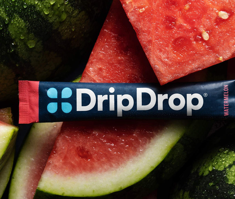 A DripDrop Watermelon flavor stick pack laid horizontally across a pile of fresh watermelon slices and rinds. The pack is dark blue with white DripDrop branding, and light blue four-dot logo.