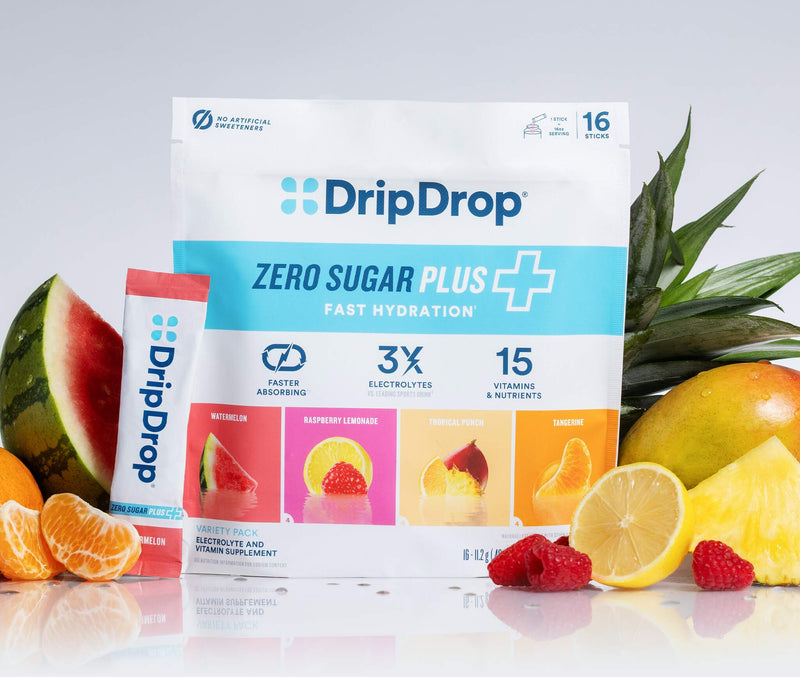 A bag and tube of DripDrop Zero Sugar Plus fast hydration drink mix are centered on a white reflective surface, surrounded by fruit slices and whole fruits including pineapple, watermelon, orange, and raspberries.
