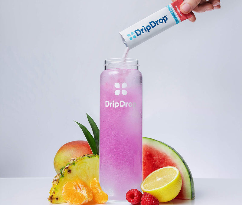 A hand pours a pink liquid from a DripDrop tube into a tall clear bottle labeled DripDrop, surrounded by fruit slices and segments on a clean white background.