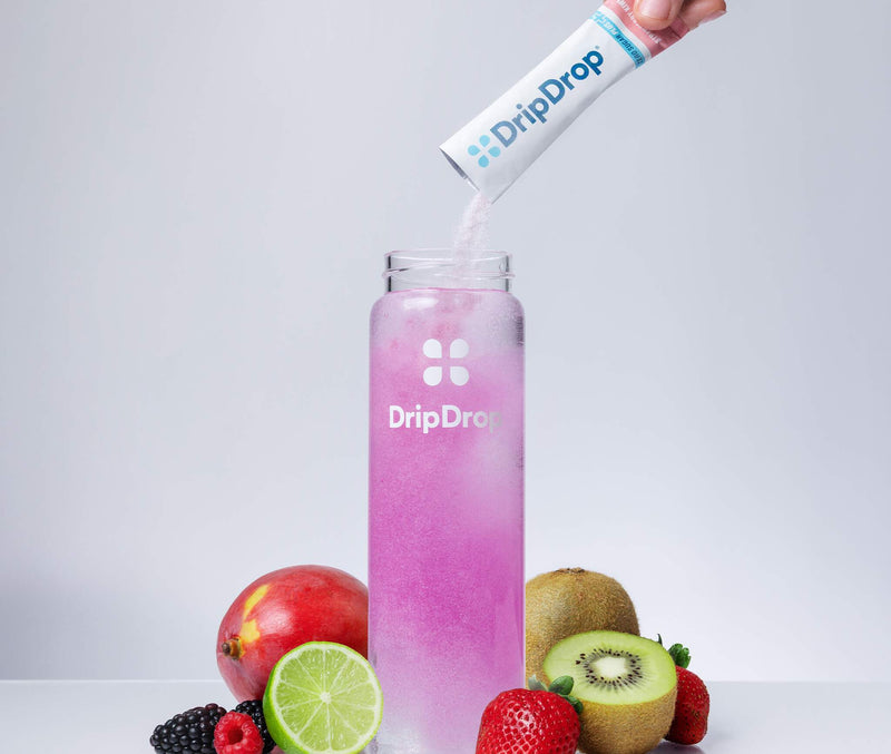 A hand pours powder from a DripDrop packet into a clear bottle filled with pink liquid, surrounded by fresh fruits including strawberries, kiwi, lime, pomegranate, and blackberries on a light gray background.