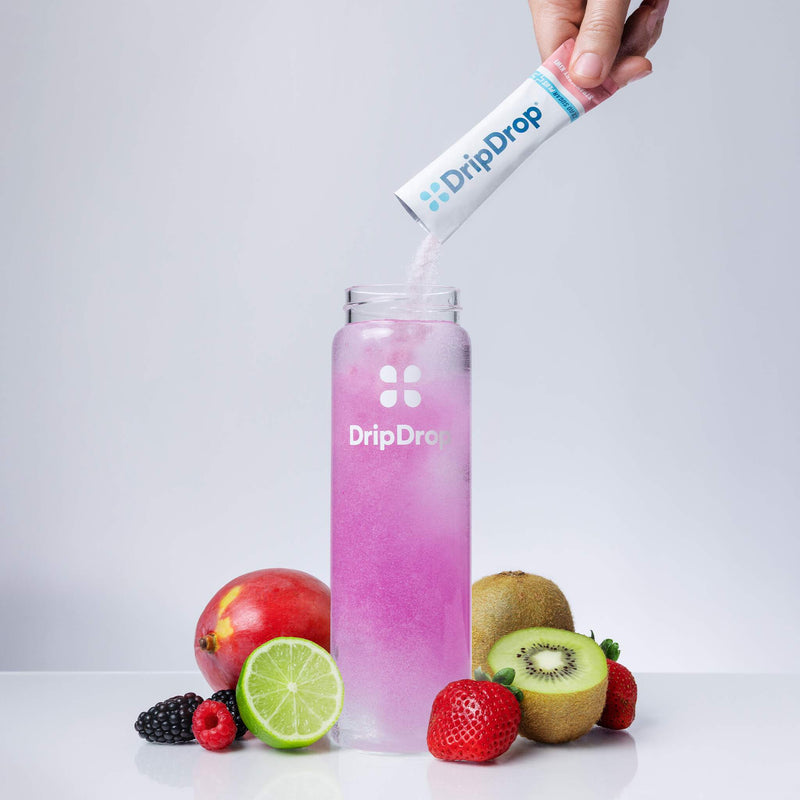 A hand pours powder from a DripDrop packet into a clear bottle filled with pink liquid, surrounded by fresh fruits including strawberries, kiwi, lime, pomegranate, and blackberries on a light gray background.