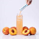 A hand pours DripDrop powder from a white and blue sachet into a clear bottle filled with peach-colored liquid, surrounded by whole and sliced peaches on a clean white background.