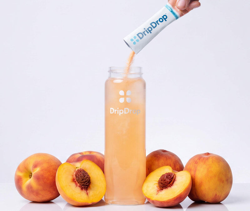 A hand pours DripDrop powder from a white and blue sachet into a clear bottle filled with peach-colored liquid, surrounded by whole and sliced peaches on a clean white background.