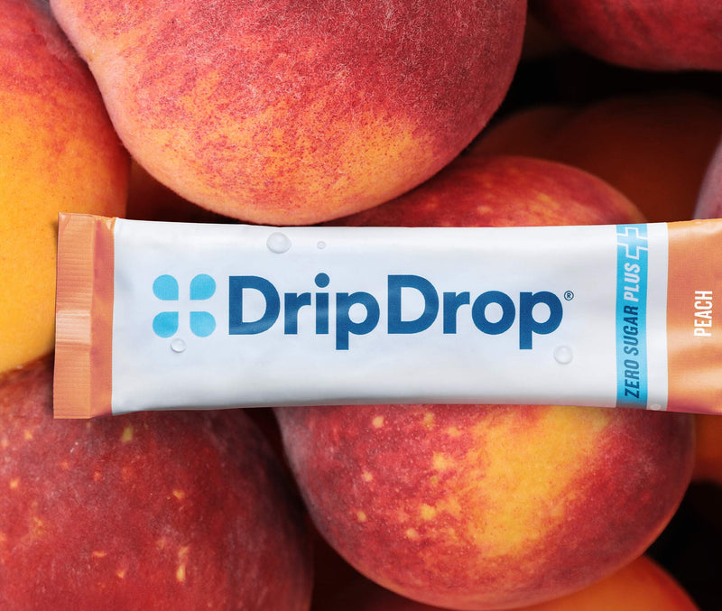 A DripDrop Zero Sugar Plus Peach flavor stick pack placed horizontally across a pile of fresh peaches.