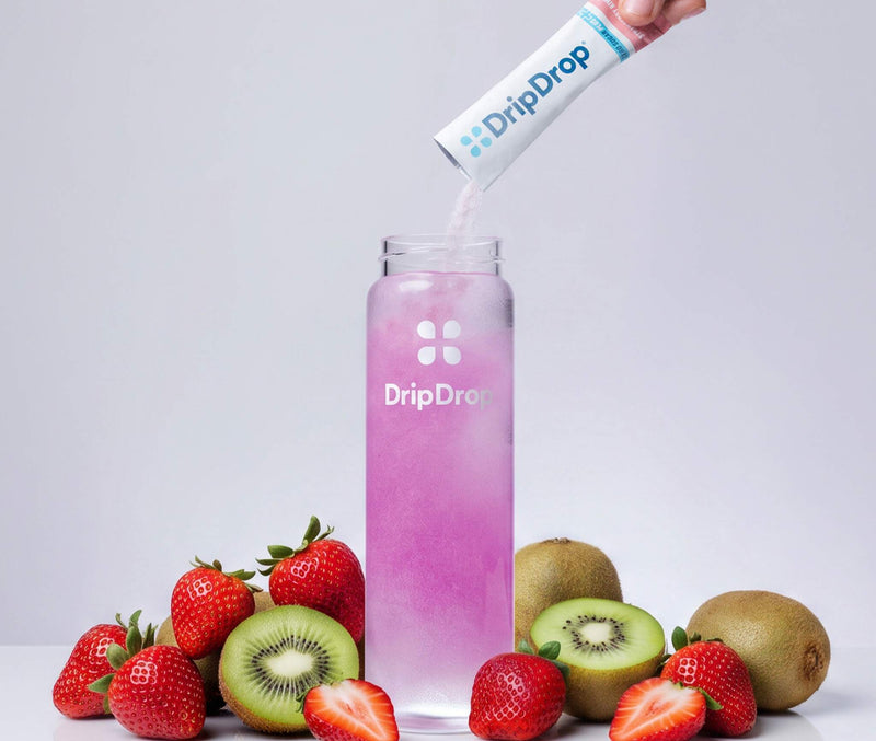 A hand pours DripDrop powder from a white sachet into a clear bottle filled with pink liquid, surrounded by fresh strawberries, kiwi slices, and whole kiwi on a light gray background.
