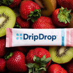 A DripDrop Zero Sugar Plus Strawberry drink mix pouch laid flat on a bed of fresh strawberries and sliced kiwi. The white and pink pouch features the blue DripDrop logo and text indicating zero sugar plus strawberry kiwi flavor.