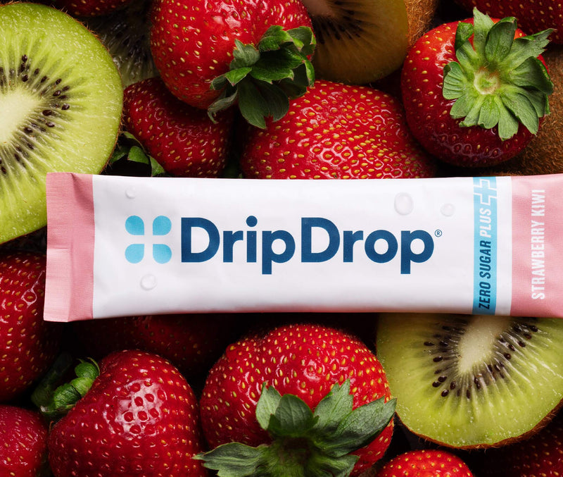 A DripDrop Zero Sugar Plus Strawberry drink mix pouch laid flat on a bed of fresh strawberries and sliced kiwi. The white and pink pouch features the blue DripDrop logo and text indicating zero sugar plus strawberry kiwi flavor.