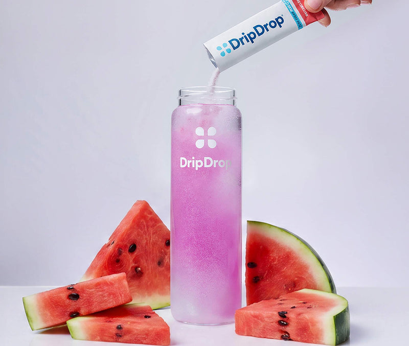 A hand pours DripDrop powder from a white and blue sachet into a clear bottle filled with pink liquid, surrounded by watermelon slices on a clean white background.