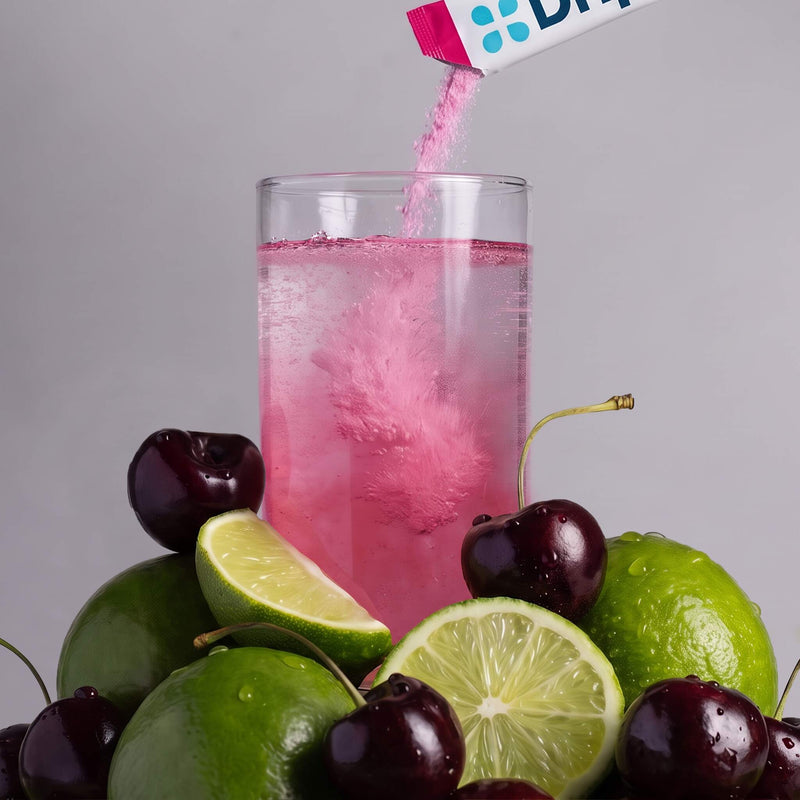 A clear glass filled with a pink drink, with a pink powder product being poured into it from a small sleeve. The glass is surrounded by green limes and dark cherries on a light gray studio background.