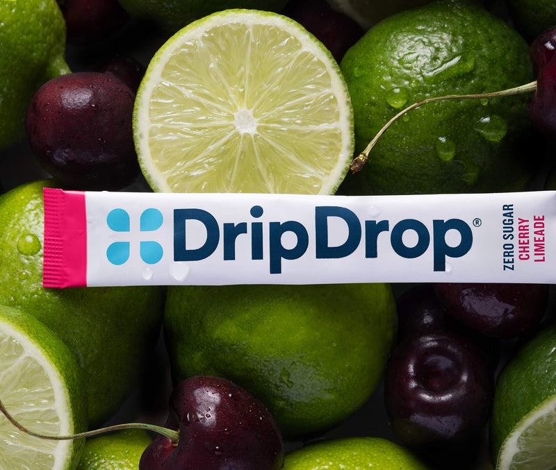 A DripDrop Zero Sugar Cherry Limeade flavor stick pack laid horizontally across a close-up arrangement of fresh limes and cherries.
