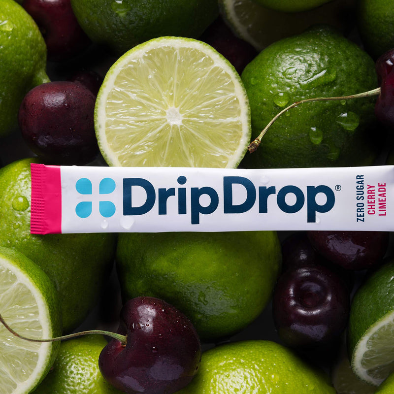 A DripDrop Zero Sugar Cherry Limeade flavor stick pack laid horizontally across a close-up arrangement of fresh limes and cherries.