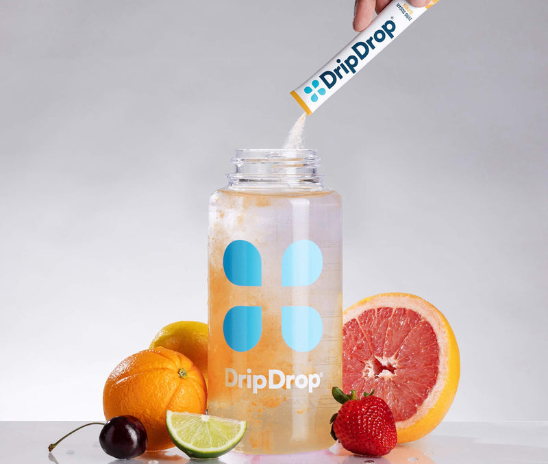 A clear plastic water bottle with blue and white DripDrop branding sits on a reflective surface, with a hand pouring powder from a DripDrop stick pack into the bottle. Fresh citrus fruits, a grapefruit half, a lime wedge, and a strawberry are arranged around the bottle against a light gray studio background.