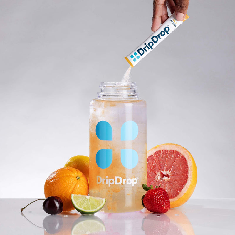 A clear plastic water bottle with blue and white DripDrop branding sits on a reflective surface, with a hand pouring powder from a DripDrop stick pack into the bottle. Fresh citrus fruits, a grapefruit half, a lime wedge, and a strawberry are arranged around the bottle against a light gray studio background.