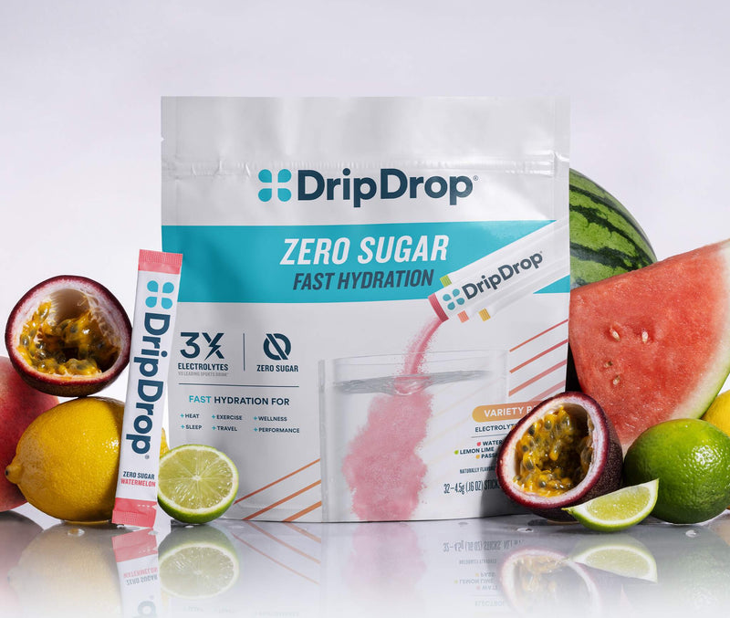 A stand-up pouch of DripDrop Zero Sugar fast hydration powder stands on a reflective white surface, surrounded by fresh fruit slices and halves including watermelon, lemon, lime, and passion fruit. A single DripDrop stick pack is placed to the left of the pouch.
