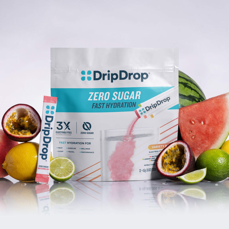 A stand-up pouch of DripDrop Zero Sugar fast hydration powder stands on a reflective white surface, surrounded by fresh fruit slices and halves including watermelon, lemon, lime, and passion fruit. A single DripDrop stick pack is placed to the left of the pouch.