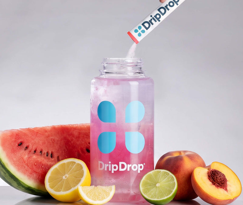 A clear plastic bottle with a pink drink inside and blue DripDrop logo graphics, with a hand pouring a DripDrop powder stick into the bottle. Watermelon, lemon, lime, and peach slices are arranged around the bottle on a light gray background.