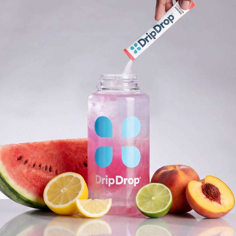 A clear plastic bottle with a pink drink inside and blue DripDrop logo graphics, with a hand pouring a DripDrop powder stick into the bottle. Watermelon, lemon, lime, and peach slices are arranged around the bottle on a light gray background.