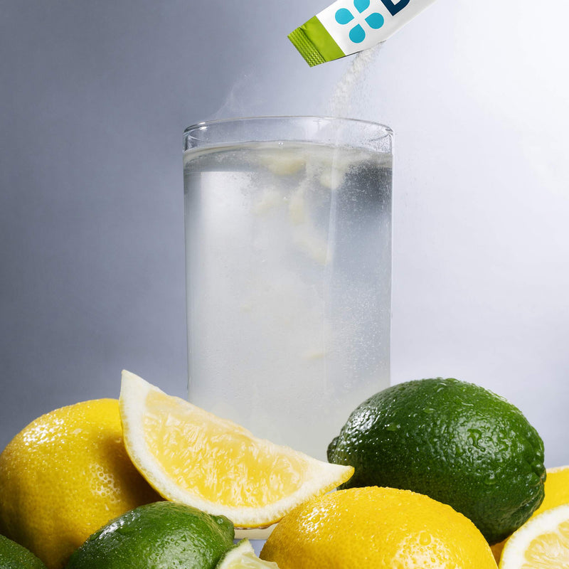 A clear glass of water with a white powder being poured from a green and white packet, surrounded by fresh lemons and limes on a gray studio background.