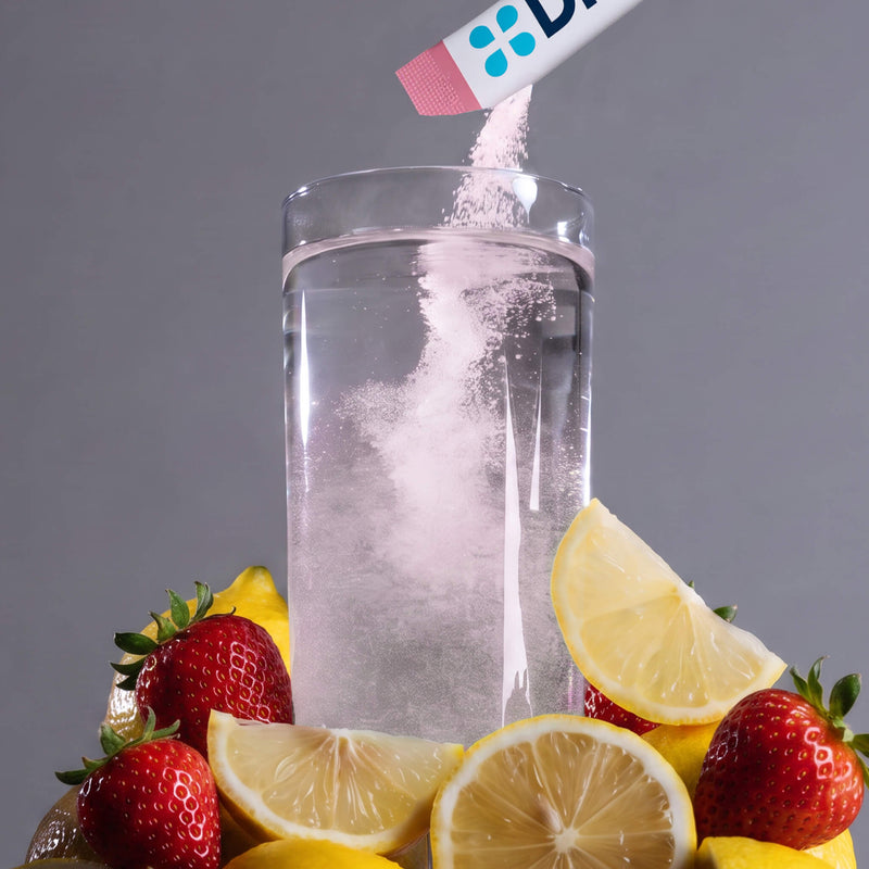 A clear glass tumbler filled with clear liquid, with a white powder being poured into it from a white tube with blue dots. The glass is surrounded by fresh strawberries and lemon slices on a gray background.