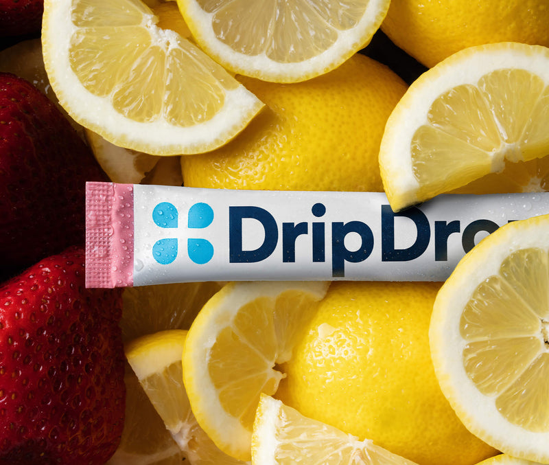 A DripDrop drink mix stick pack placed horizontally across a close-up arrangement of fresh lemon slices and strawberries. The package is white with blue branding and a pink end cap, surrounded by bright yellow lemons and red strawberries.