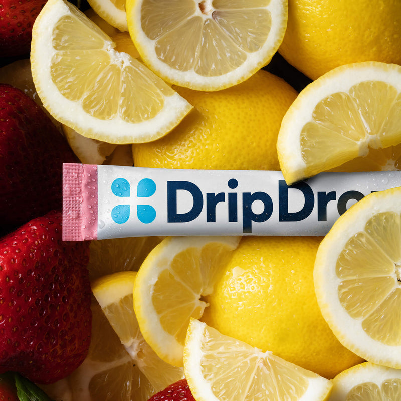 A DripDrop drink mix stick pack placed horizontally across a close-up arrangement of fresh lemon slices and strawberries. The package is white with blue branding and a pink end cap, surrounded by bright yellow lemons and red strawberries.