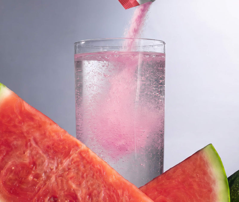 A clear glass filled with a clear bubbly liquid, with pink powder being poured into it from a small packet. Large watermelon slices frame the foreground and background against a soft gray studio backdrop.