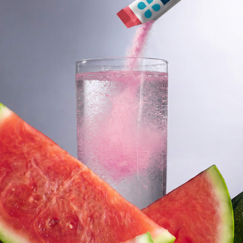 A clear glass filled with a clear bubbly liquid, with pink powder being poured into it from a small packet. Large watermelon slices frame the foreground and background against a soft gray studio backdrop.