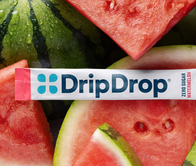 A DripDrop watermelon flavor zero sugar stick pack laid horizontally across a close-up background of sliced watermelon and a whole watermelon.
