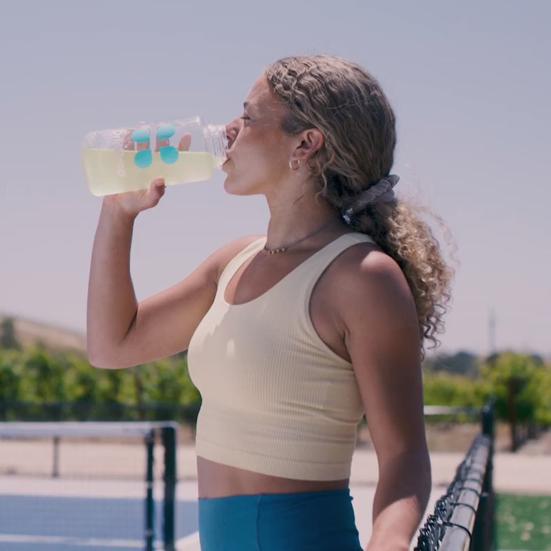 Woman drinking yellow liquid from a water bottle on a tennis court