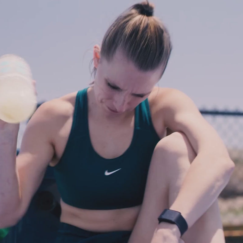 Woman shaking a water bottle filled with DripDrop and drinking it on a tennis court